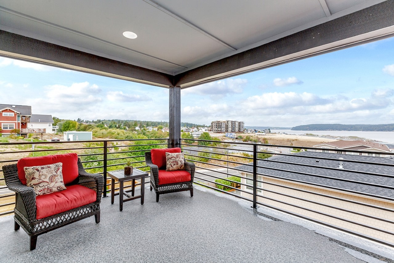 A spacious covered balcony with two red cushioned chairs overlooking a body of water and coastal properties.
