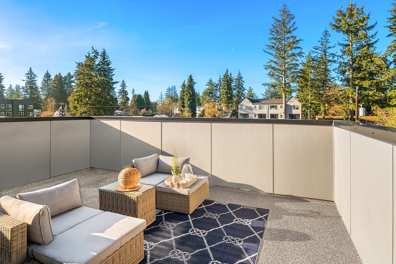 A private rooftop patio with comfortable wicker seating on a patterned rug, surrounded by tall evergreen trees.