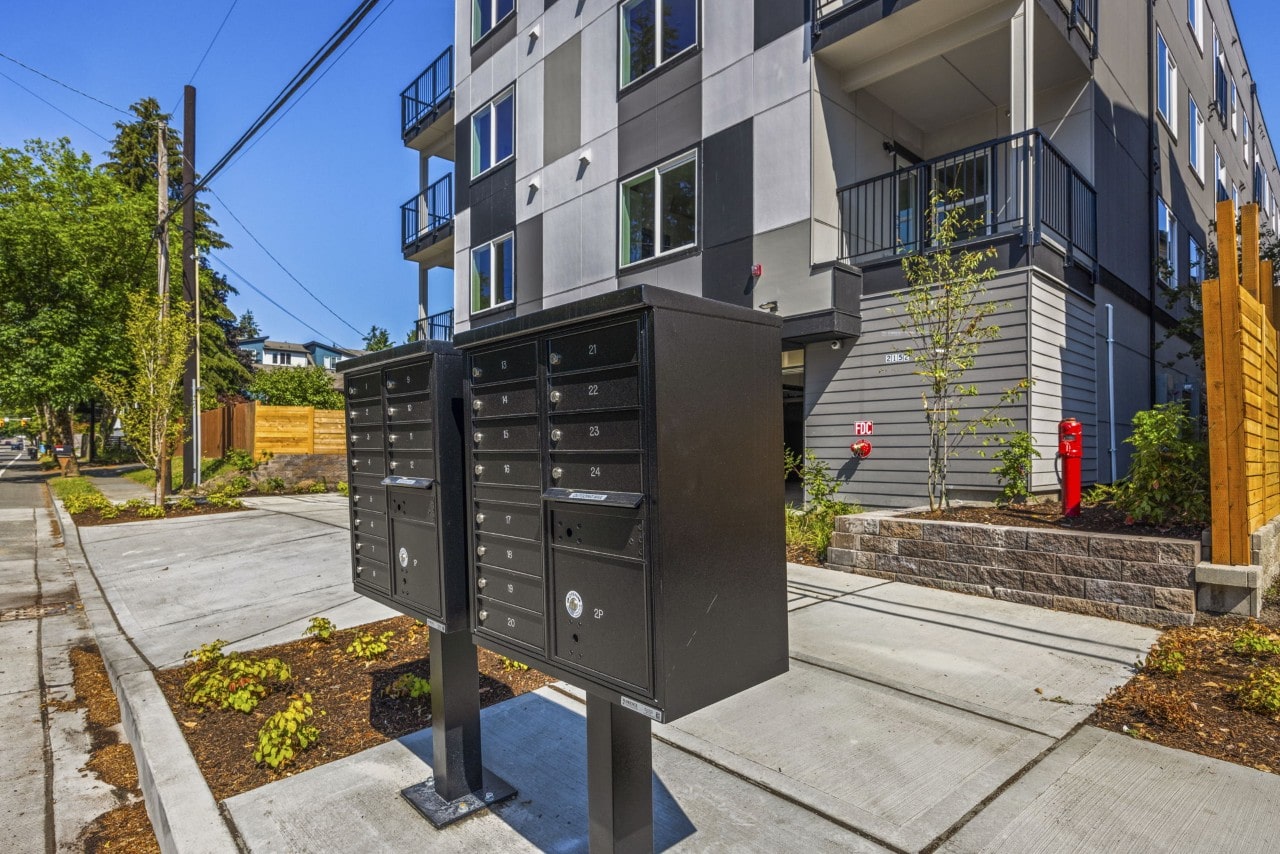 Two black community mailboxes on a sidewalk in front of a modern apartment building exterior.
