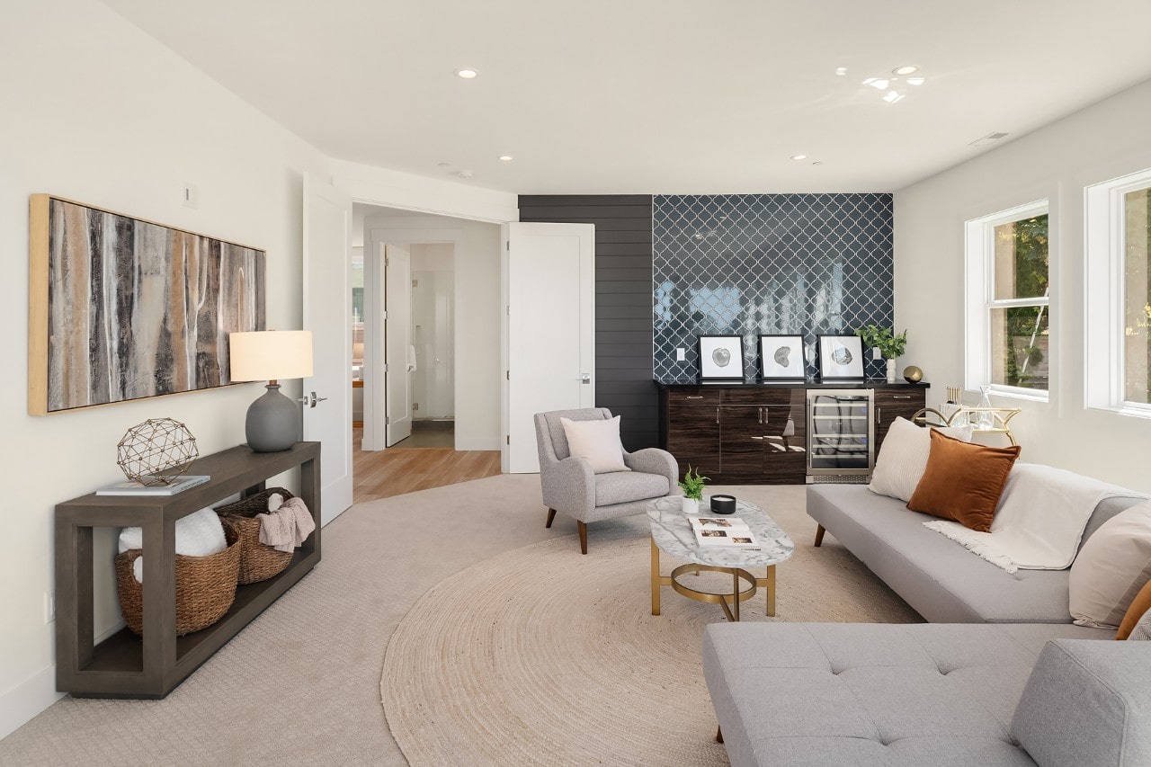 A cozy, modern lounge area with a gray sofa, a wet bar featuring a blue tile backsplash, and a wine fridge.