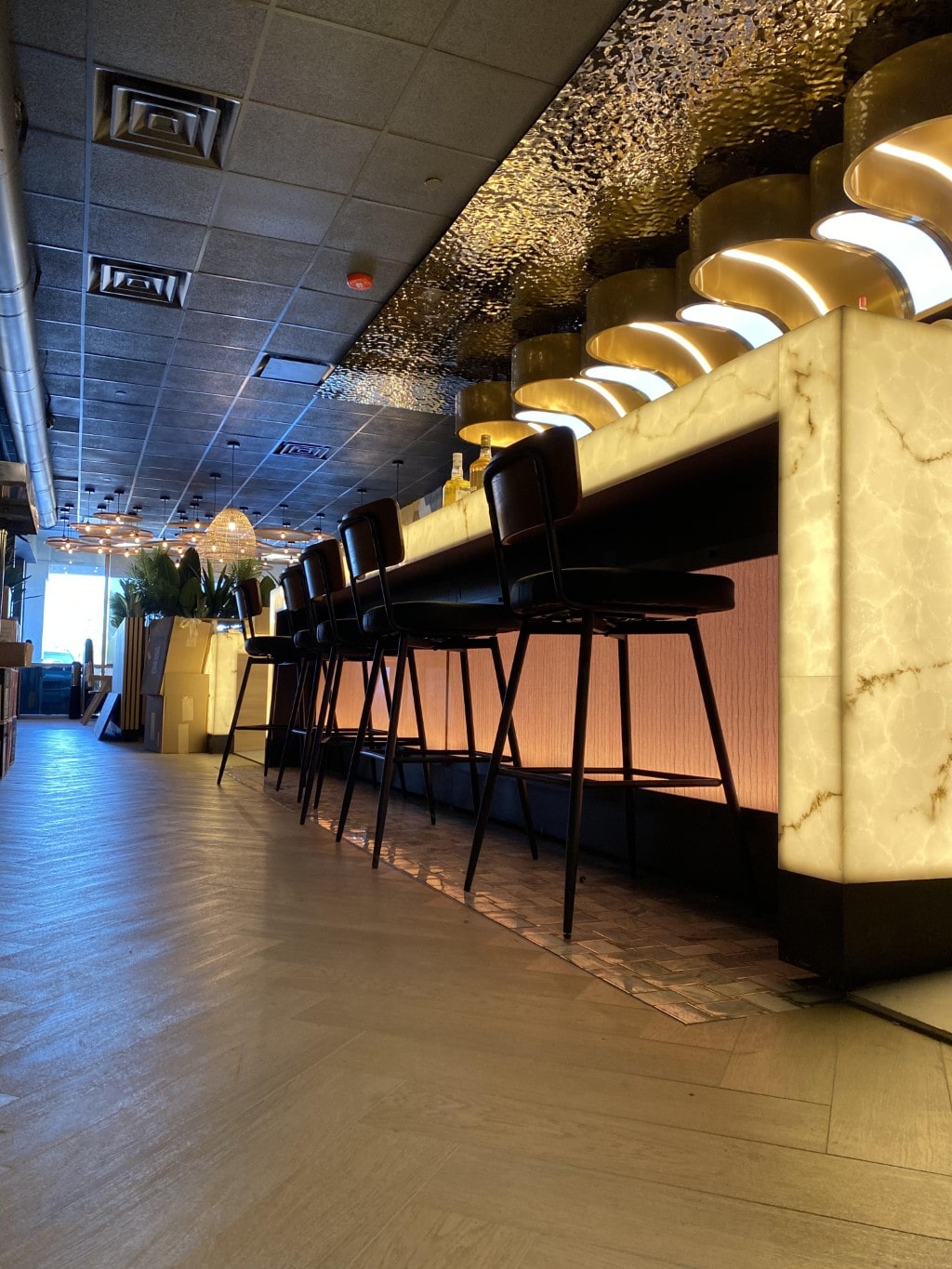 A side view of the modern bar at Basil Bistro, featuring an illuminated white marble countertop, black high-top chairs, and unique gold overhead lighting fixtures.