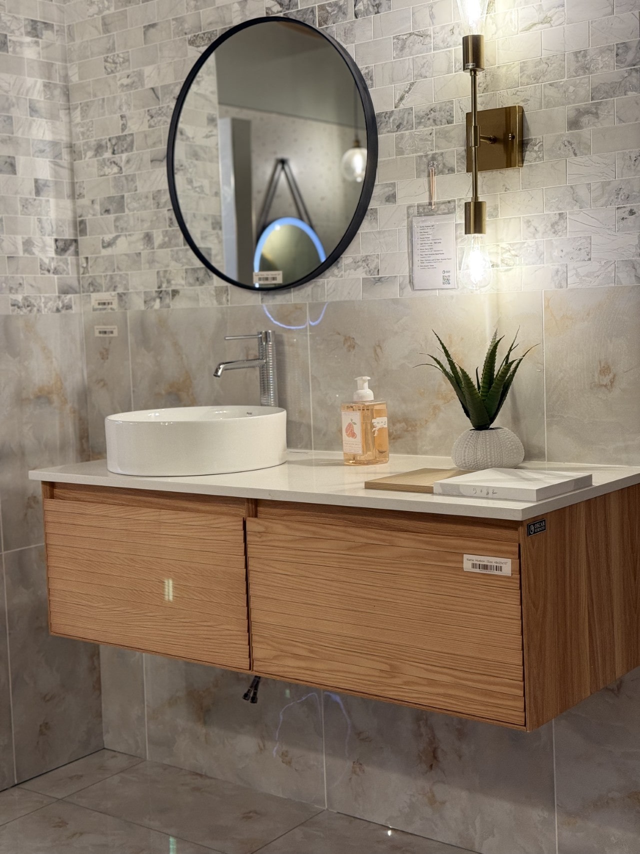 A modern bathroom vignette featuring a floating wood vanity, a white vessel sink, a round black mirror, and a wall of grey marbled tiles.