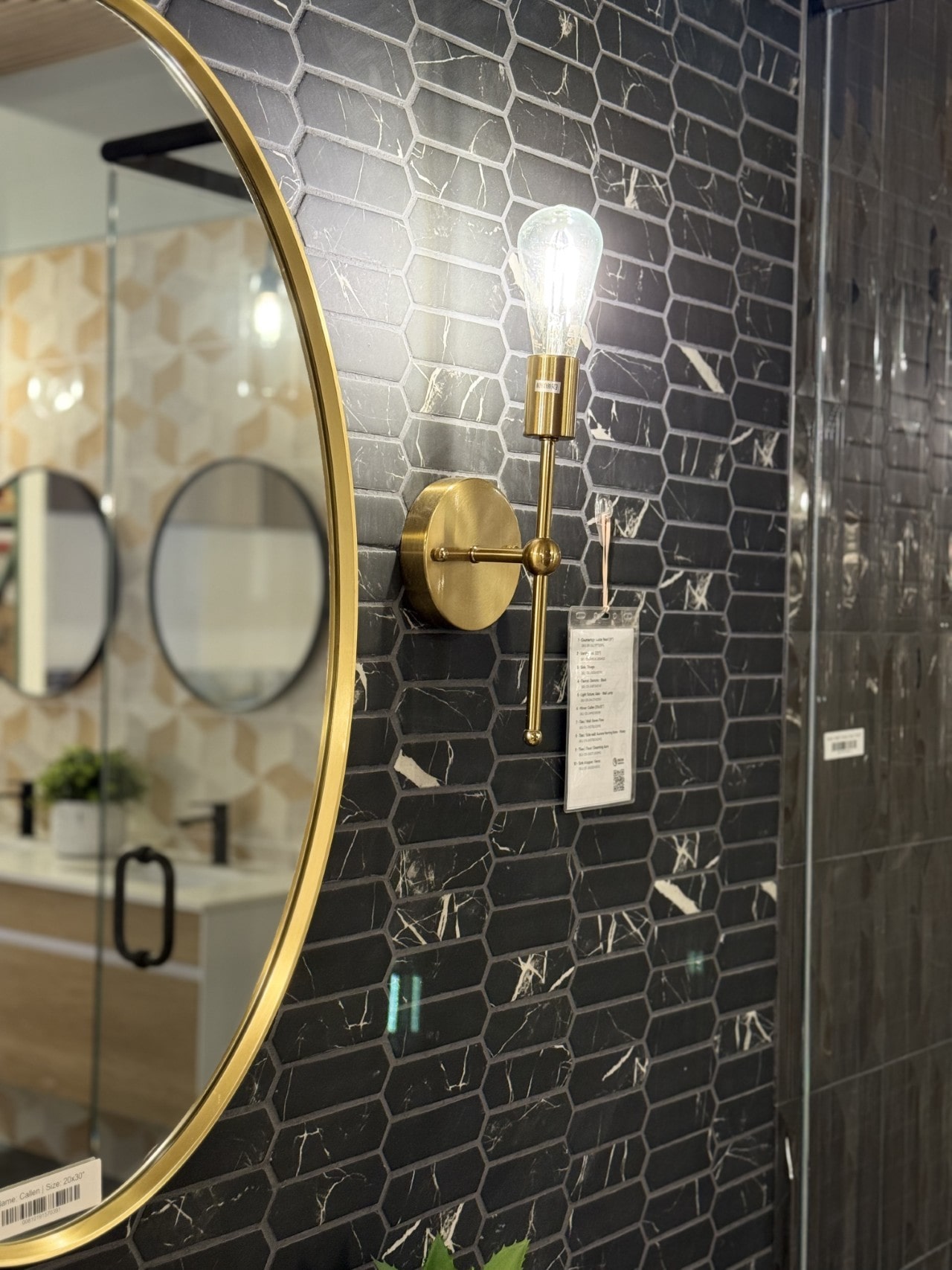 A close-up of a stylish bathroom wall with black picket-shaped tiles, a modern brass sconce, and the edge of a gold-framed circular mirror.