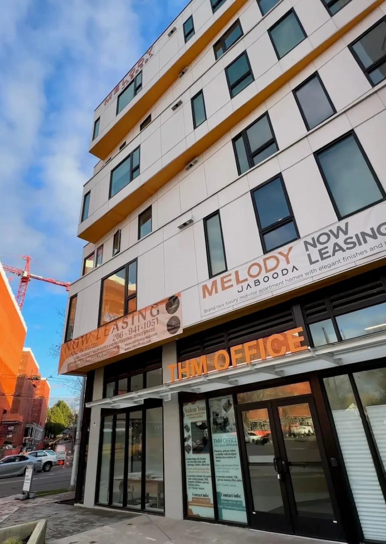 A street view looking up at the modern "Melody Jabooda" apartment building, with "Now Leasing" banners displayed on its white and yellow facade.