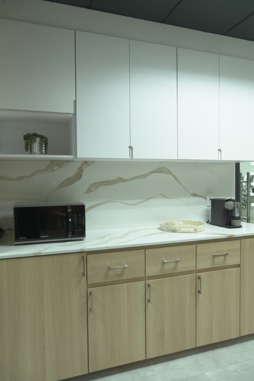 A close-up of a clean, modern kitchenette with light wood base cabinets, a white marble-look countertop and backsplash, and white upper cabinets.