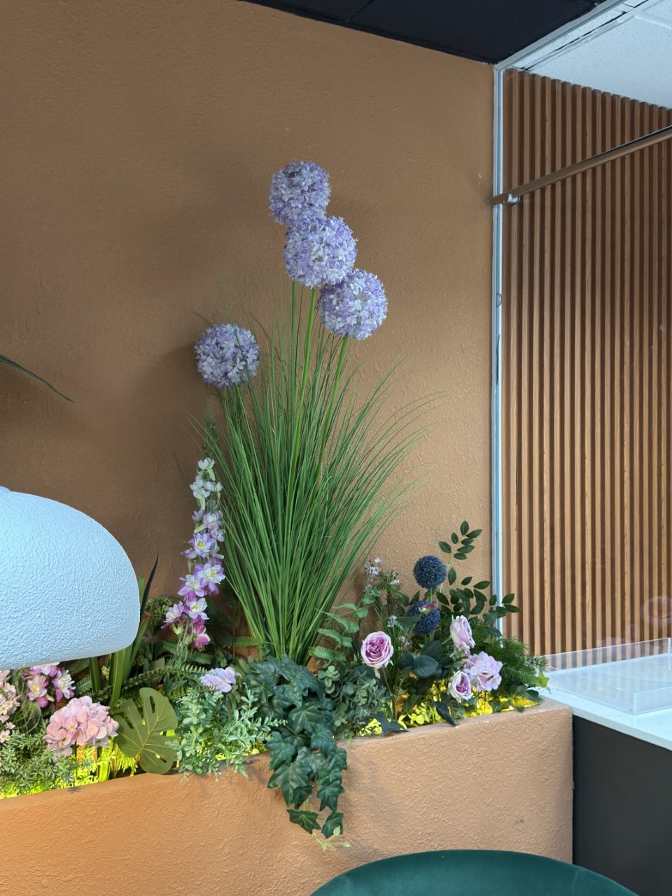 A close-up of an indoor planter with lush artificial greenery and tall, spherical purple flowers against a textured terracotta-colored wall.