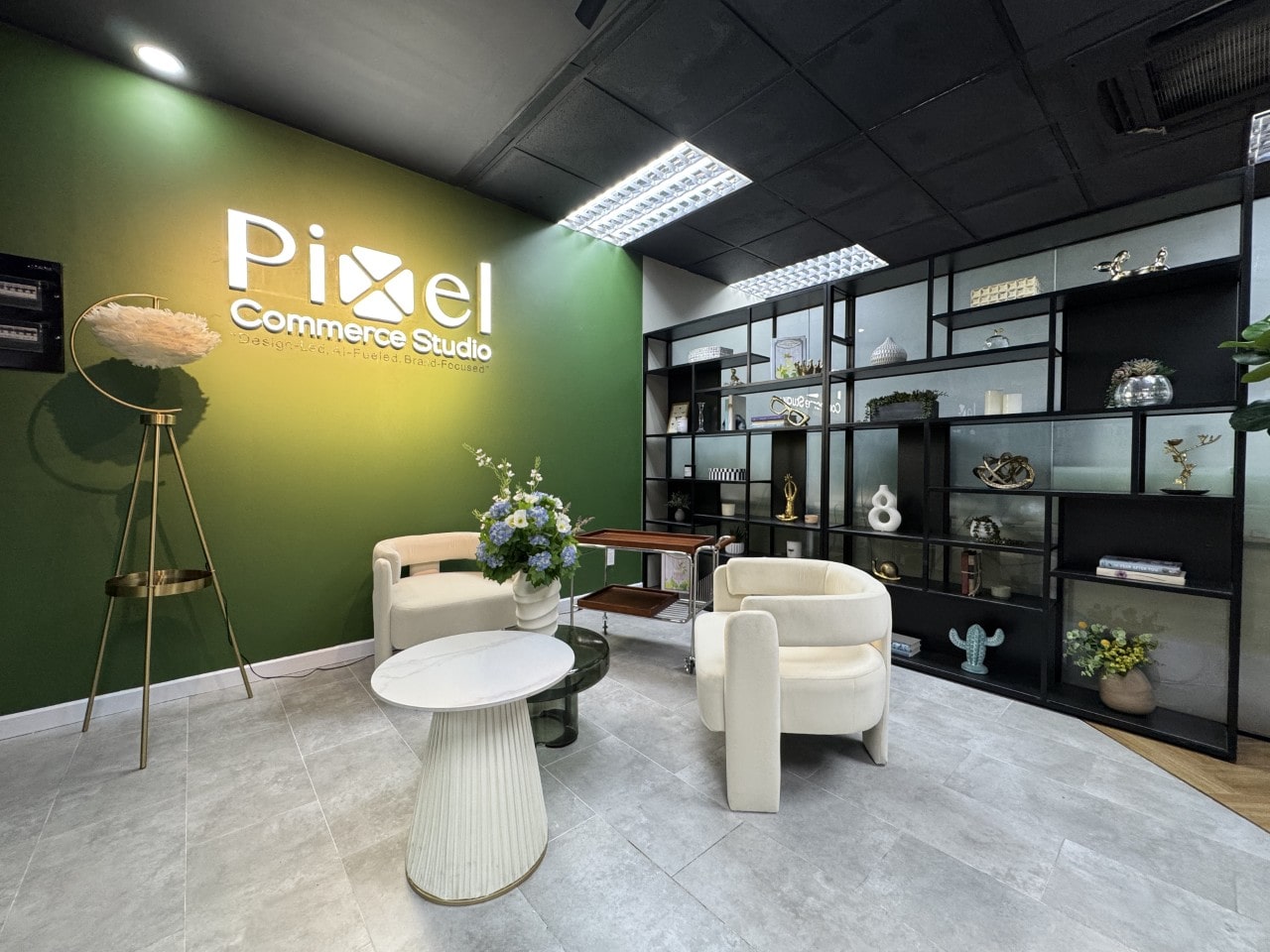 The reception area of Pixel Commerce Studio with a large backlit logo on a green wall, cream armchairs, and a large black shelving unit.