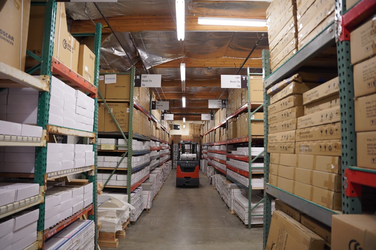 An aisle in the Oscar Surfaces warehouse with tall shelves stacked high with boxes of inventory and a forklift in the distance