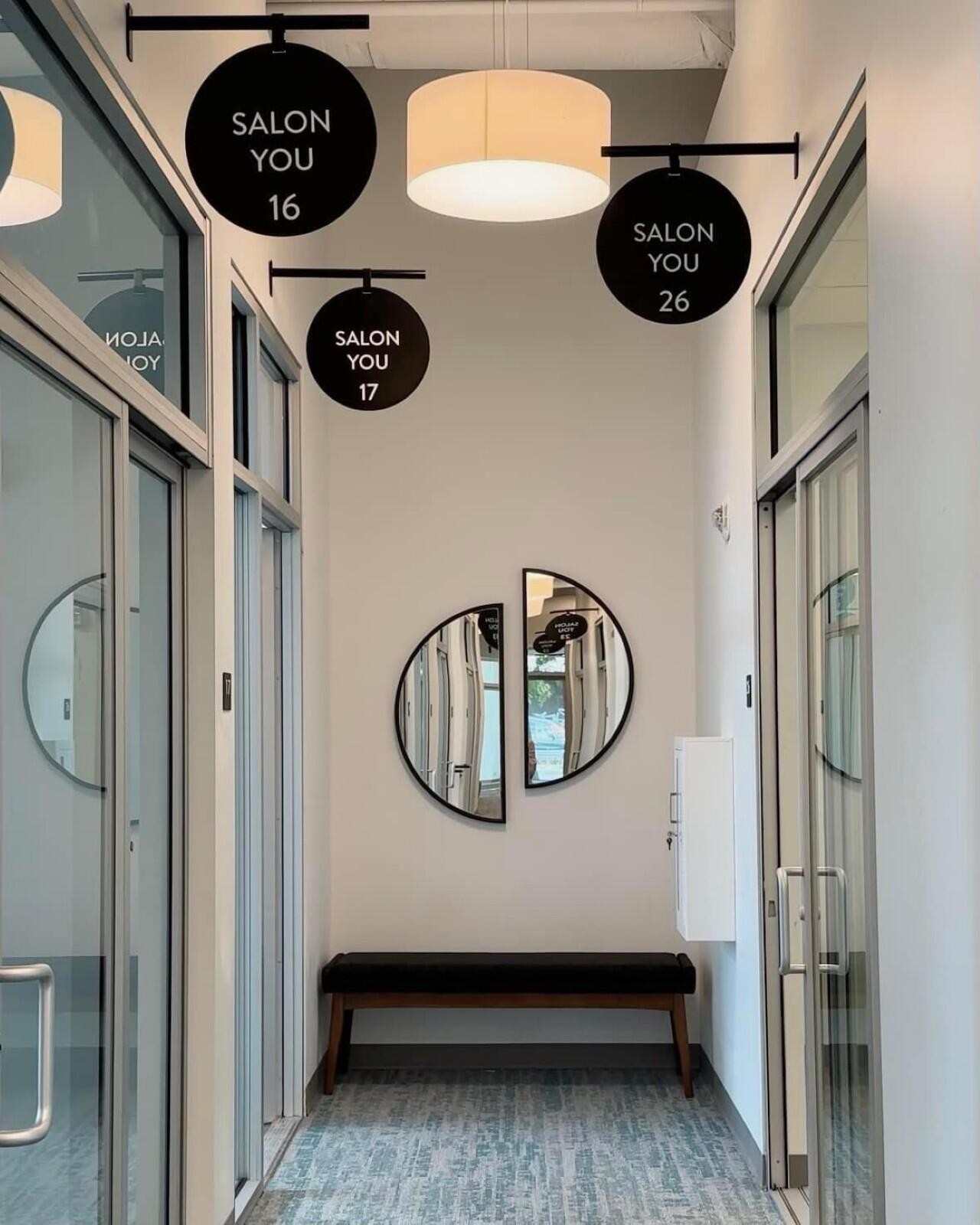 A hallway at Salon You with numbered circular signs for different suites, a bench, and a unique split-circle mirror on the wall.