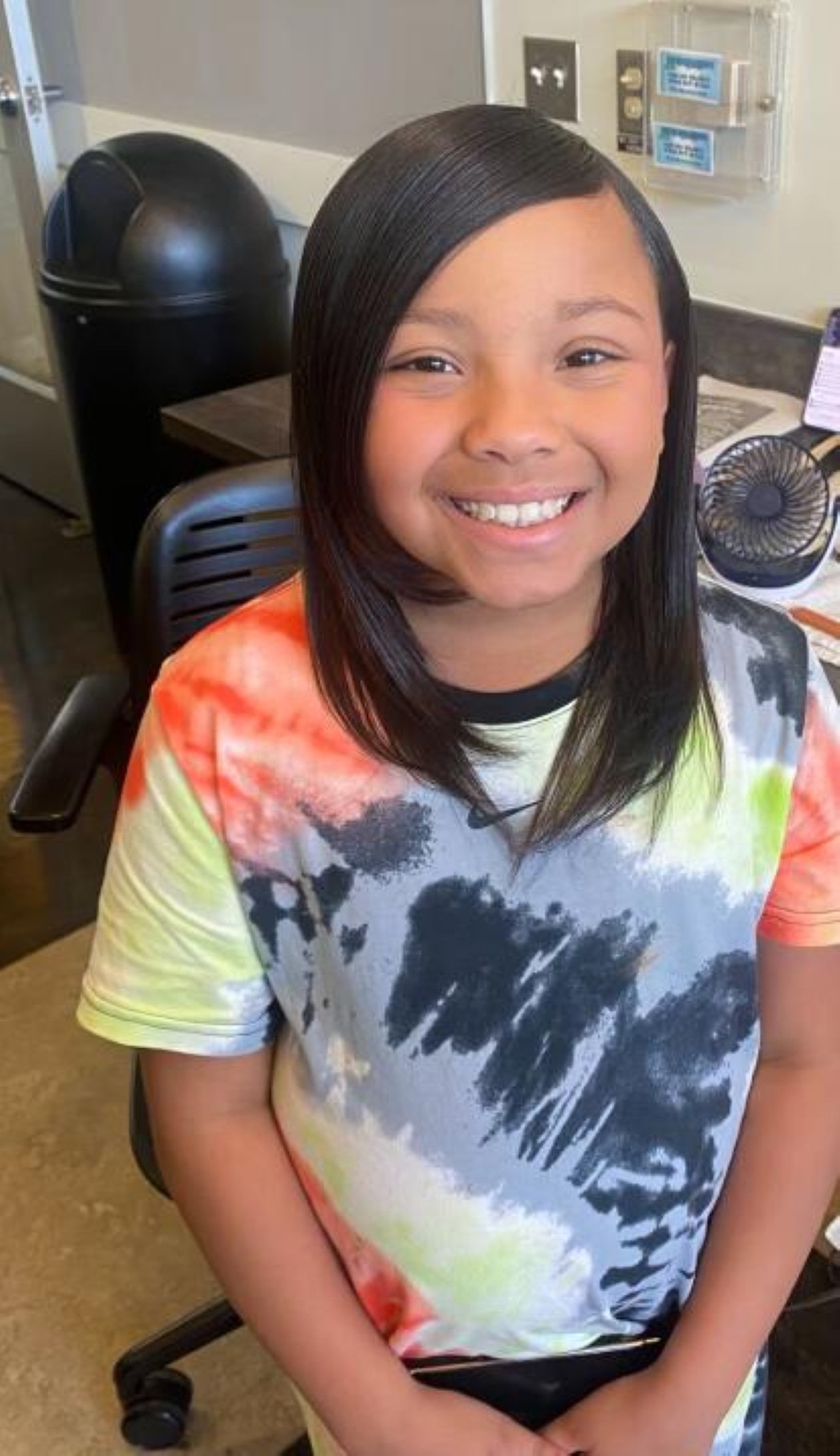 A smiling young girl with a sleek, straight side-part hairstyle sitting in a salon chair.