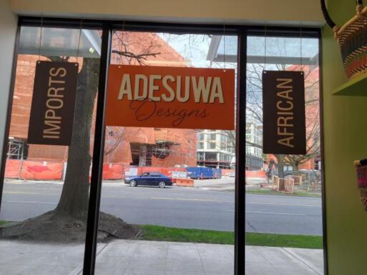 View from a storefront window with "ADESUWA Designs" and "AFRICAN IMPORTS" signs hanging, looking out onto a street with construction.