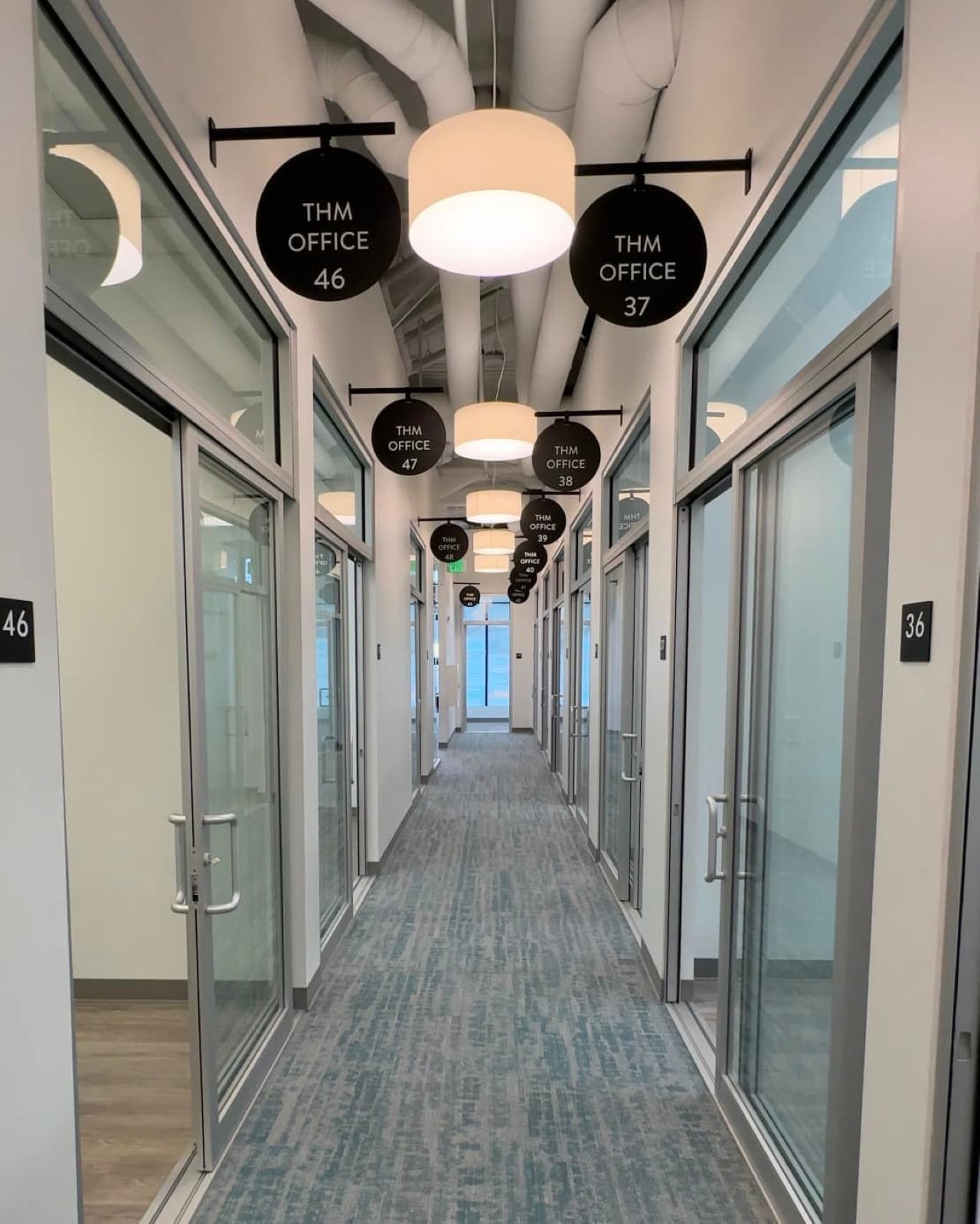 A long, modern hallway inside THM Office with glass-walled office doors on either side and circular signs indicating office numbers hanging from the ceiling.