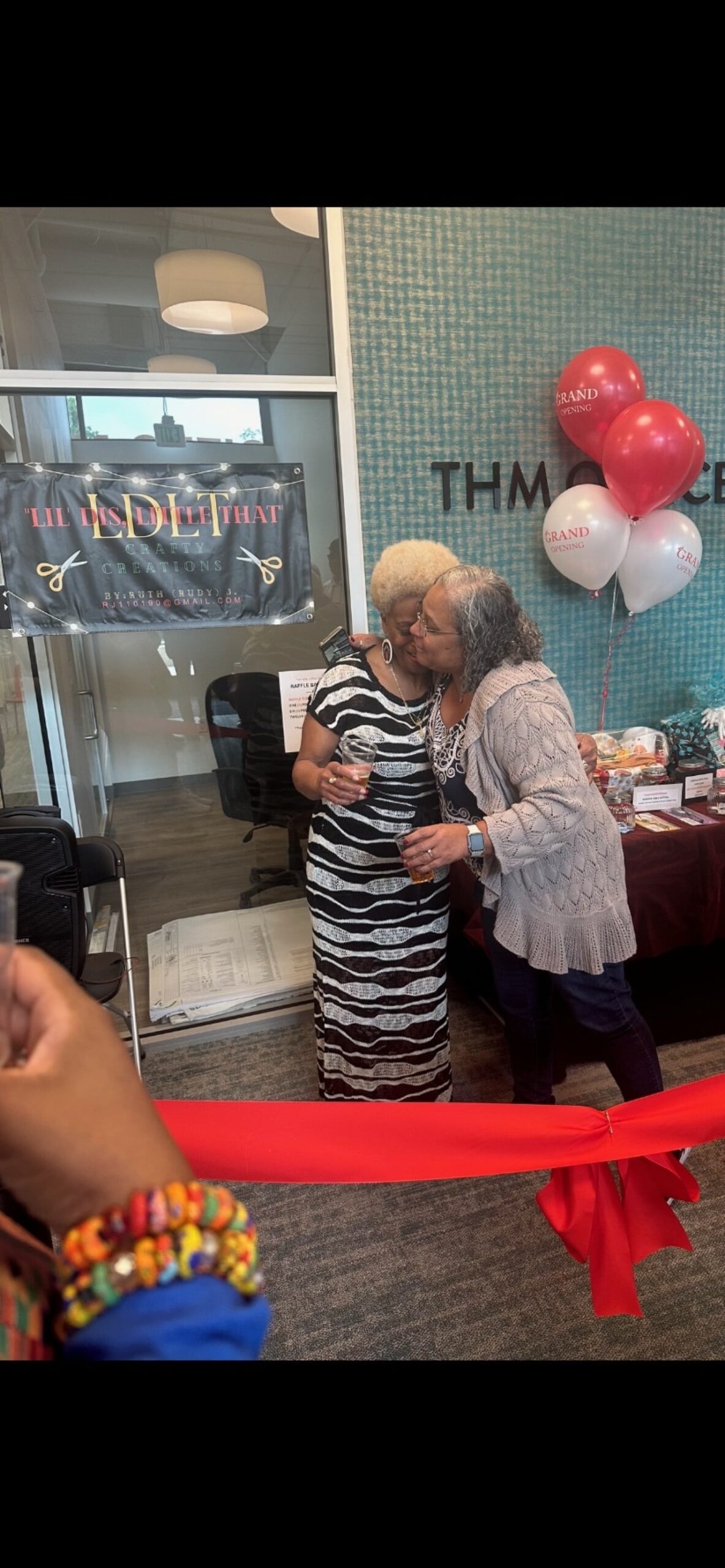  Two women hugging warmly during a celebration event at THM Office, with a red ribbon in the foreground and a business banner in the background.