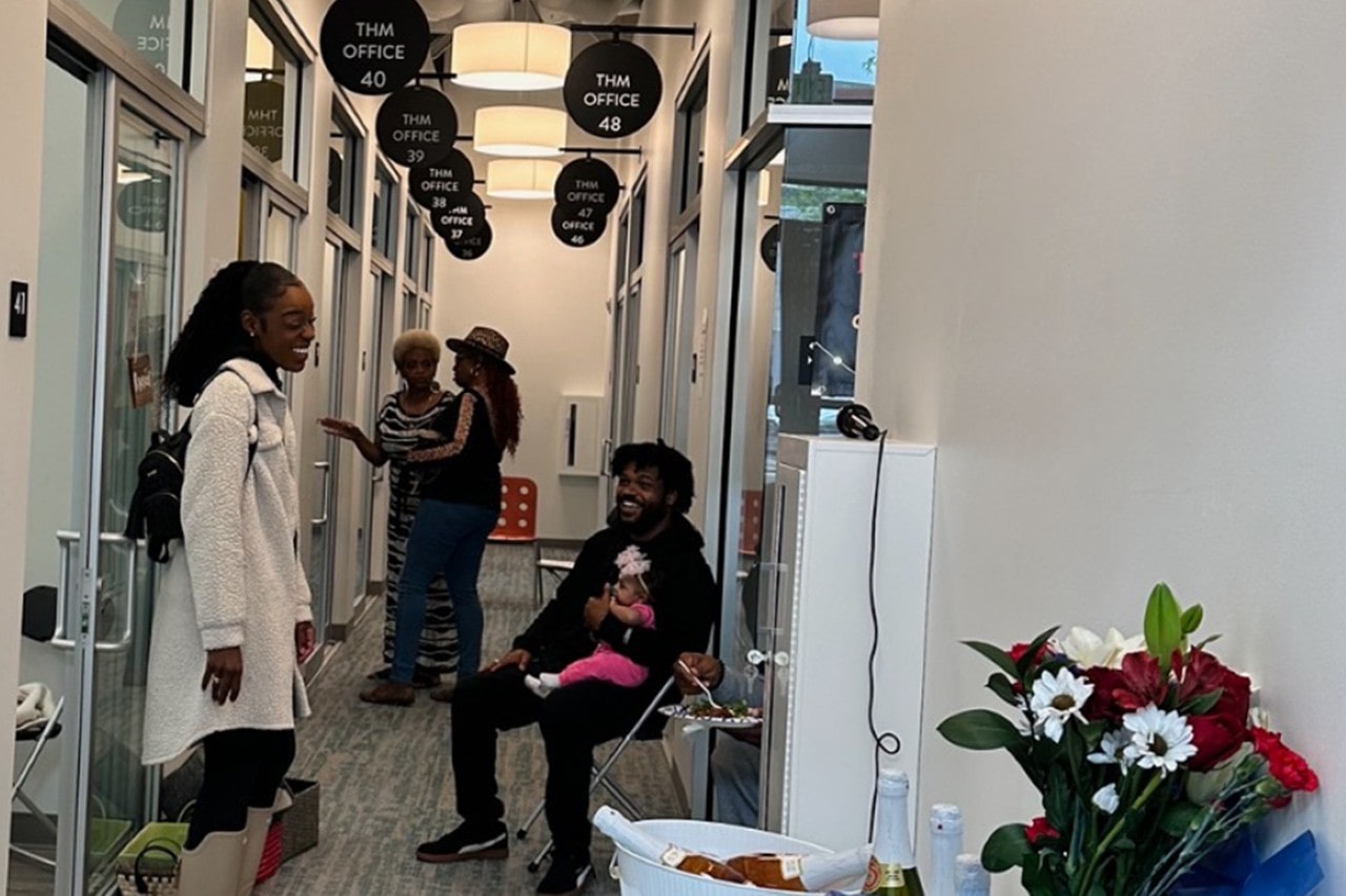 A candid photo inside the THM Office hallway during an event, with people talking, a man holding a baby, and a celebratory table with flowers and champagne in the foreground.