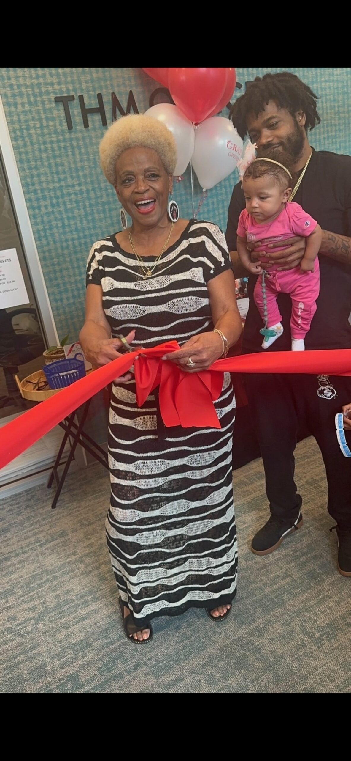 An older woman joyfully cuts a red ribbon at a grand opening ceremony for THM Office, with a man holding a baby and balloons behind her.
